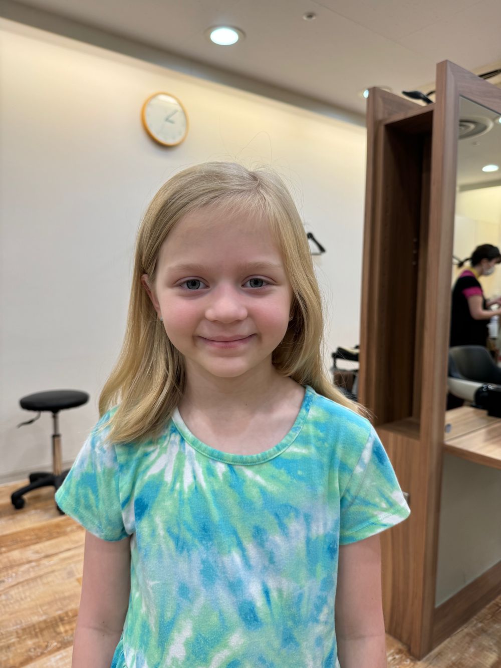 A young girl in a tie-dye shirt smiles after getting a haircut, standing in a salon with a clock and salon chairs visible behind her.