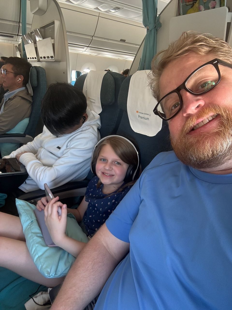 A man with glasses and a beard takes a selfie on a Vietnam Airlines flight, with a young girl wearing headphones and holding an iPad smiling beside him in the premium cabin.
