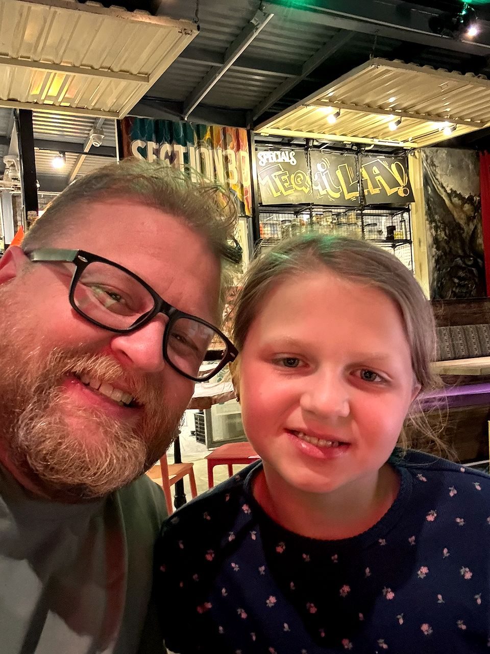 Adam leans in for a selfie with his daughter Cora inside an industrial-style Da Nang bar with a tequila specials sign painted on the wall behind them