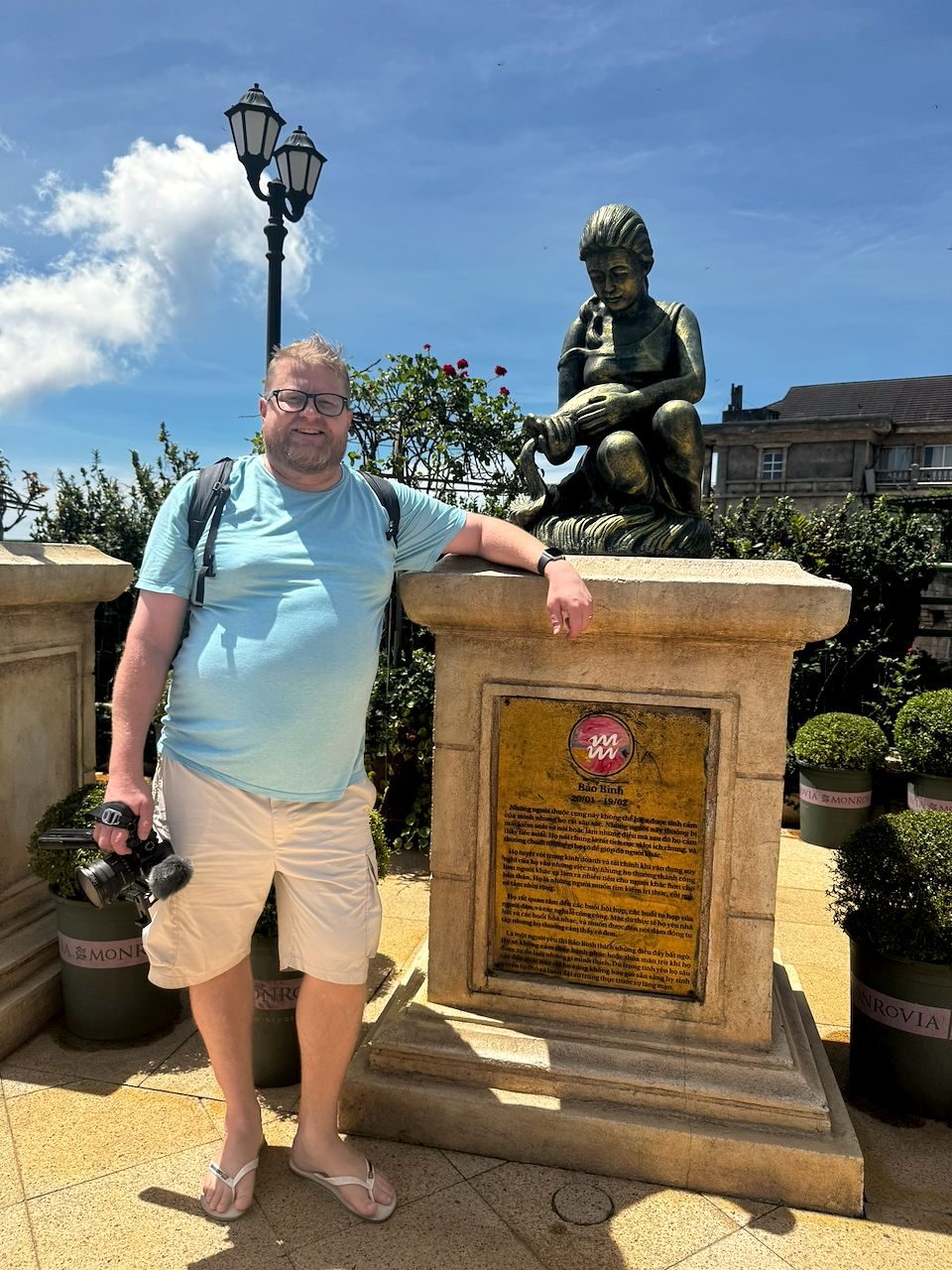 A man standing and smiling next to a bronze Aquarius zodiac statue on a stone pedestal in the flower garden at Ba Na Hills on a sunny day