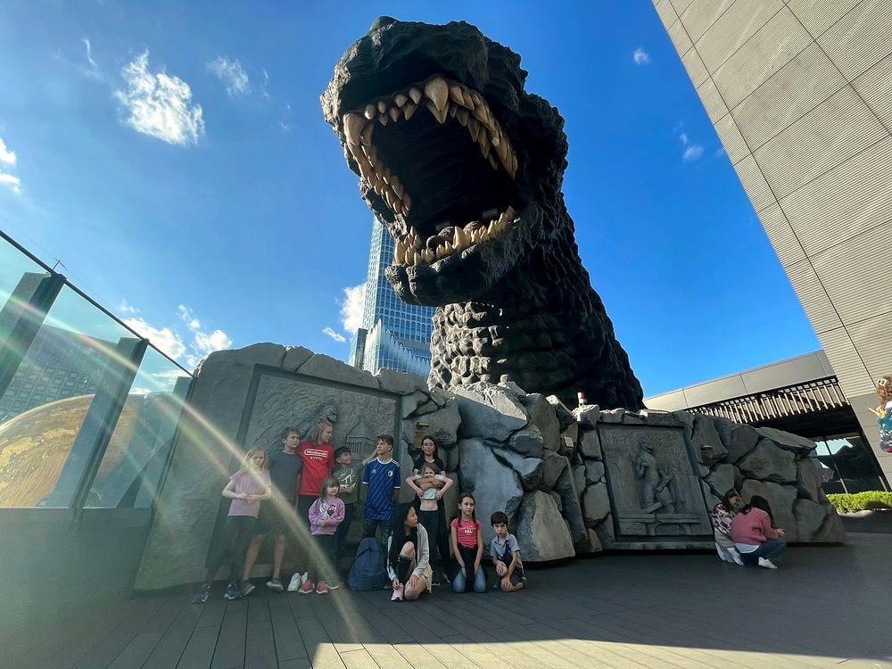 A large group of children and adults stand on an outdoor platform looking up at the enormous Godzilla head statue at Kabukicho Tower in Shinjuku against a blue sky.