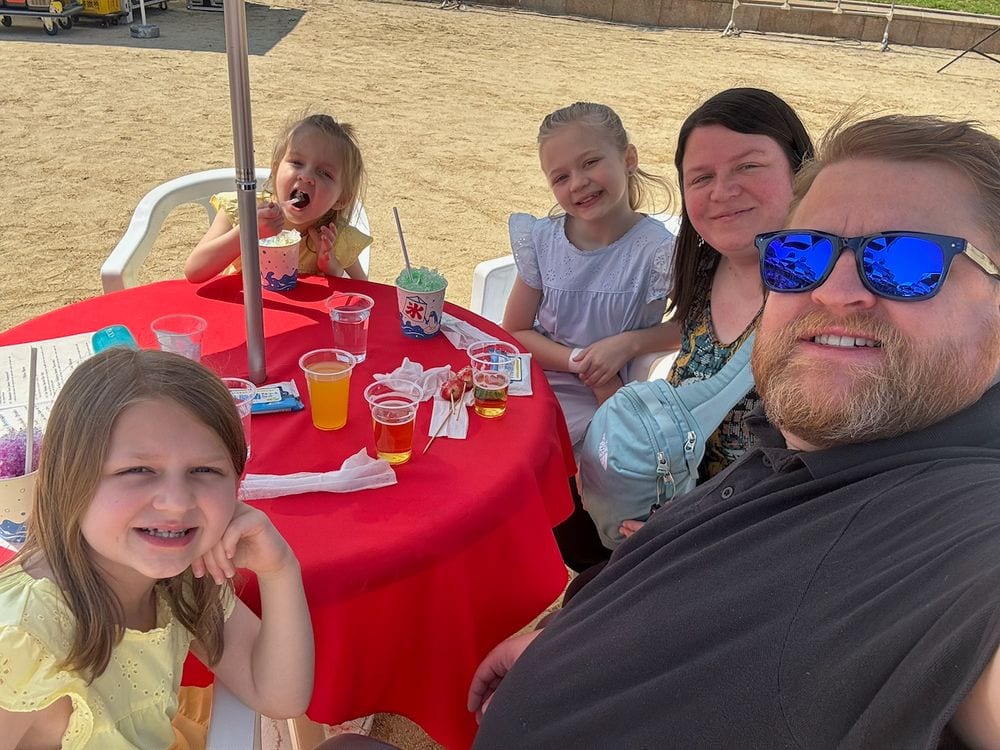 A family of five sits around a red outdoor table in Osaka, with kakigori shaved ice and juice drinks in front of them, the youngest girl eating with a spoon while the rest smile at the camera.