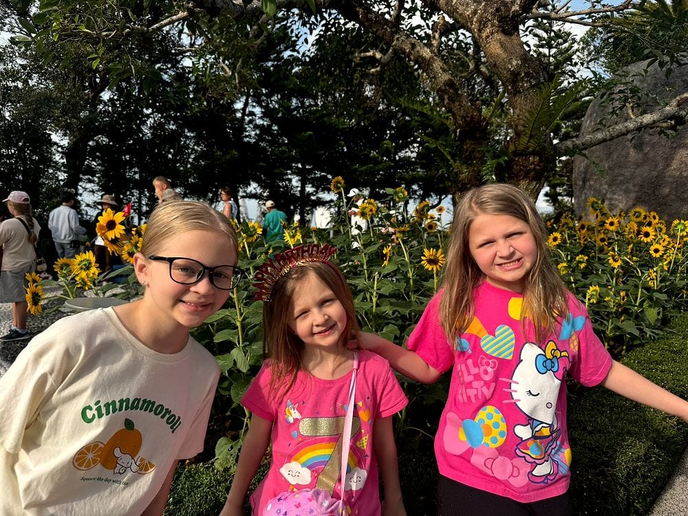 Three young girls standing and smiling in front of a field of sunflowers at Ba Na Hills, the middle girl wearing a sparkly gold Happy Birthday crown