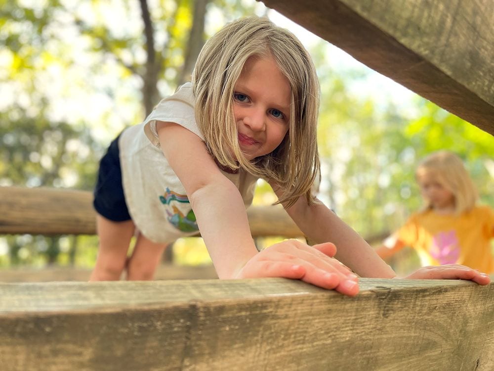 A young girl climbs a wooden beam obstacle at Heiwa no Mori Park in Ota, Tokyo, with another girl visible in the background on a rope net structure.
