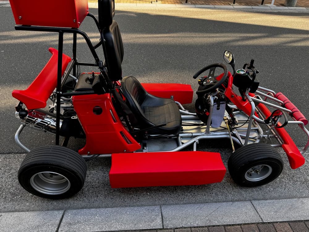 A red two-seat go-kart parked on a sidewalk in Asakusa, Tokyo, with a driver's seat, passenger seat behind it, steering wheel, and chunky black tires visible in afternoon light.