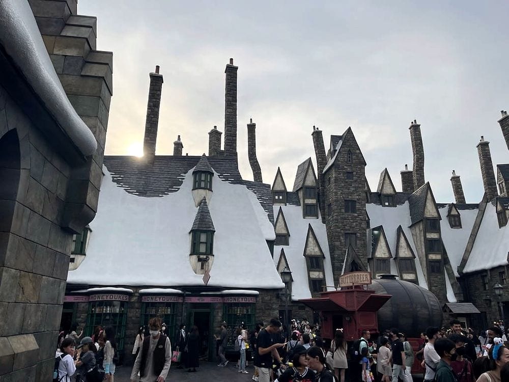 A wide shot looking up at the snow-covered rooftops of Hogsmeade village at Universal Studios Japan, with the Honeydukes storefront and a Butterbeer cart visible among a large crowd of visitors under an overcast sky.