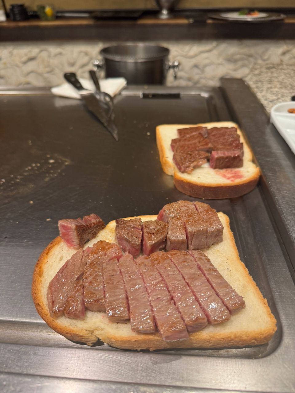 Two slices of thick white toast topped with sliced and cubed medium-rare wagyu beef resting on a teppanyaki iron griddle, with cooking tongs and a pot visible in the background.