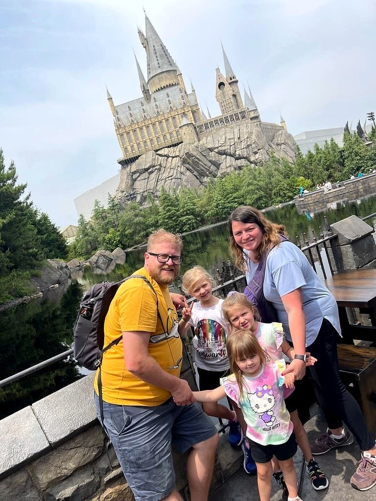 A family of five poses in front of the Hogwarts Castle replica at Universal Studios Japan, with the man in a yellow shirt on the left, a woman in a light blue shirt on the right, and three young girls between them.