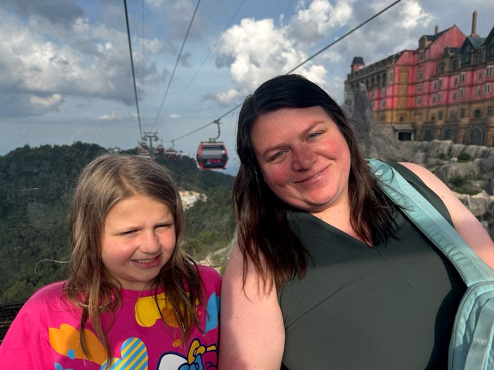 A woman and a young girl inside a cable car gondola at Ba Na Hills, with an ornate red French-style hotel and other cable cars emerging from the clouds behind them