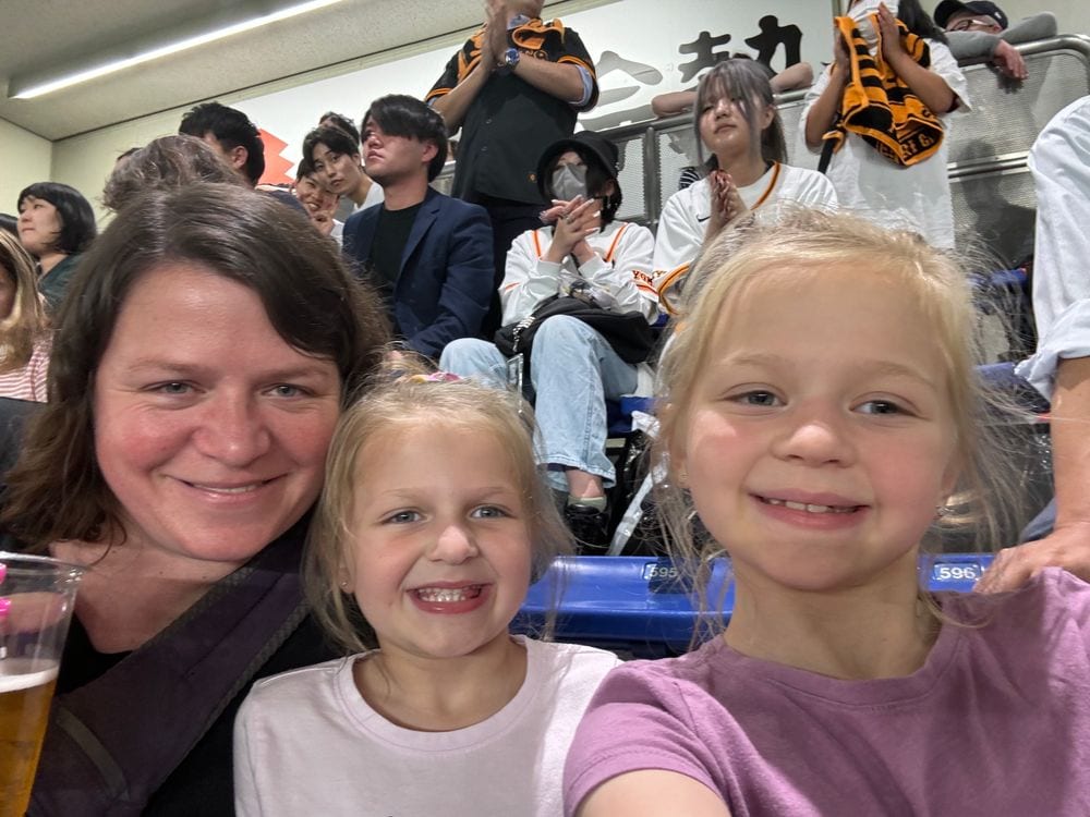 Lindsay and two young girls smile for a selfie in stadium seats at Tokyo Dome, with cheering fans visible behind them.