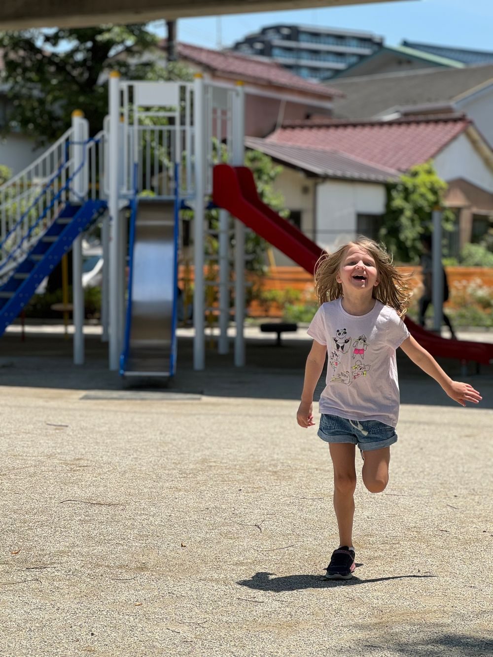 A young girl runs and laughs across a playground in Katsushika, Tokyo, with a slide and apartment buildings visible behind her.