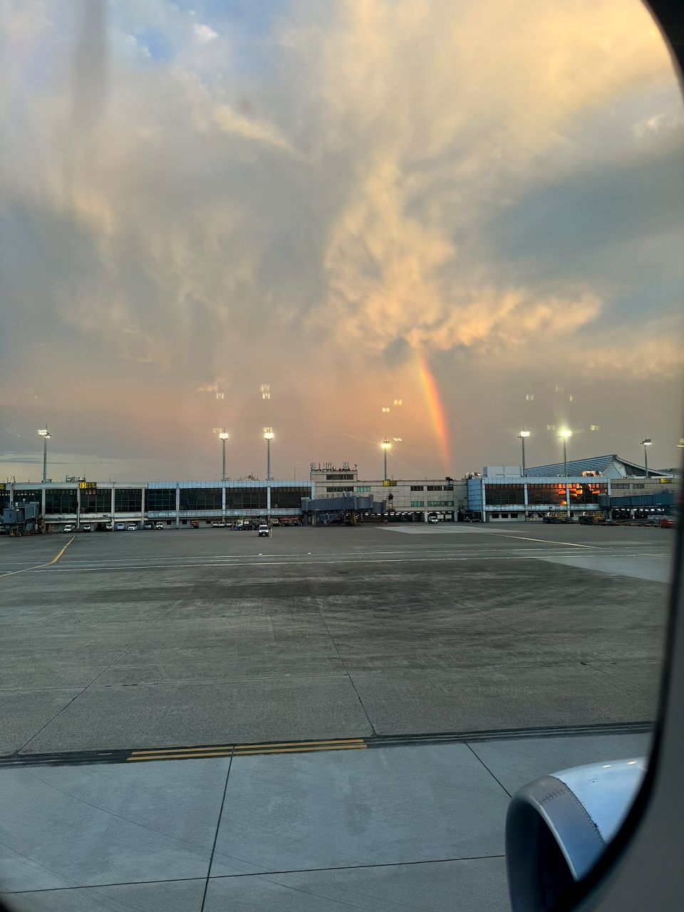 View through an airplane window of Taoyuan Airport's tarmac at dusk, with a rainbow arcing above the terminal building against a cloudy warm-toned sky, the engine nacelle visible in the lower corner.
