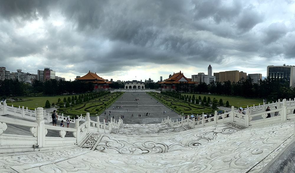 A wide panoramic shot from the top steps of Chiang Kai-shek Memorial Hall looking down across Liberty Square's manicured gardens toward the Liberty Gate and twin traditional halls, under a heavy overcast sky with storm clouds gathering over the Taipei skyline.