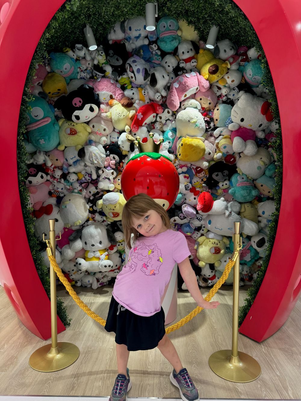 A young girl stands in front of a red apple-shaped photo arch surrounded by a wall of Sanrio character plush toys.