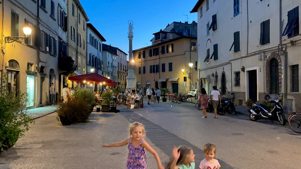 Three young girls walk and run through a quiet Italian piazza at blue hour, with warm restaurant lights and a column monument visible in the background