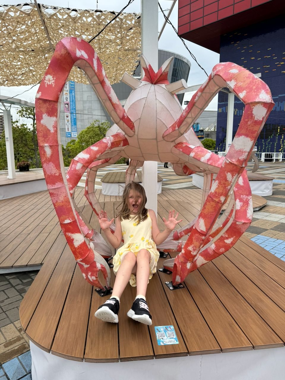 A girl in a yellow dress sits inside a large decorative spider crab sculpture on a wooden deck along the Osaka waterfront, arms raised and mouth open in an excited expression.