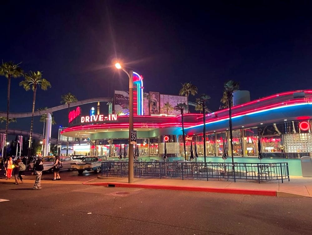 A wide night shot of Mel's Drive-In restaurant at Universal Studios Japan, its red and blue neon trim glowing against a dark sky, with palm trees, vintage cars, and scattered park guests visible in the foreground.
