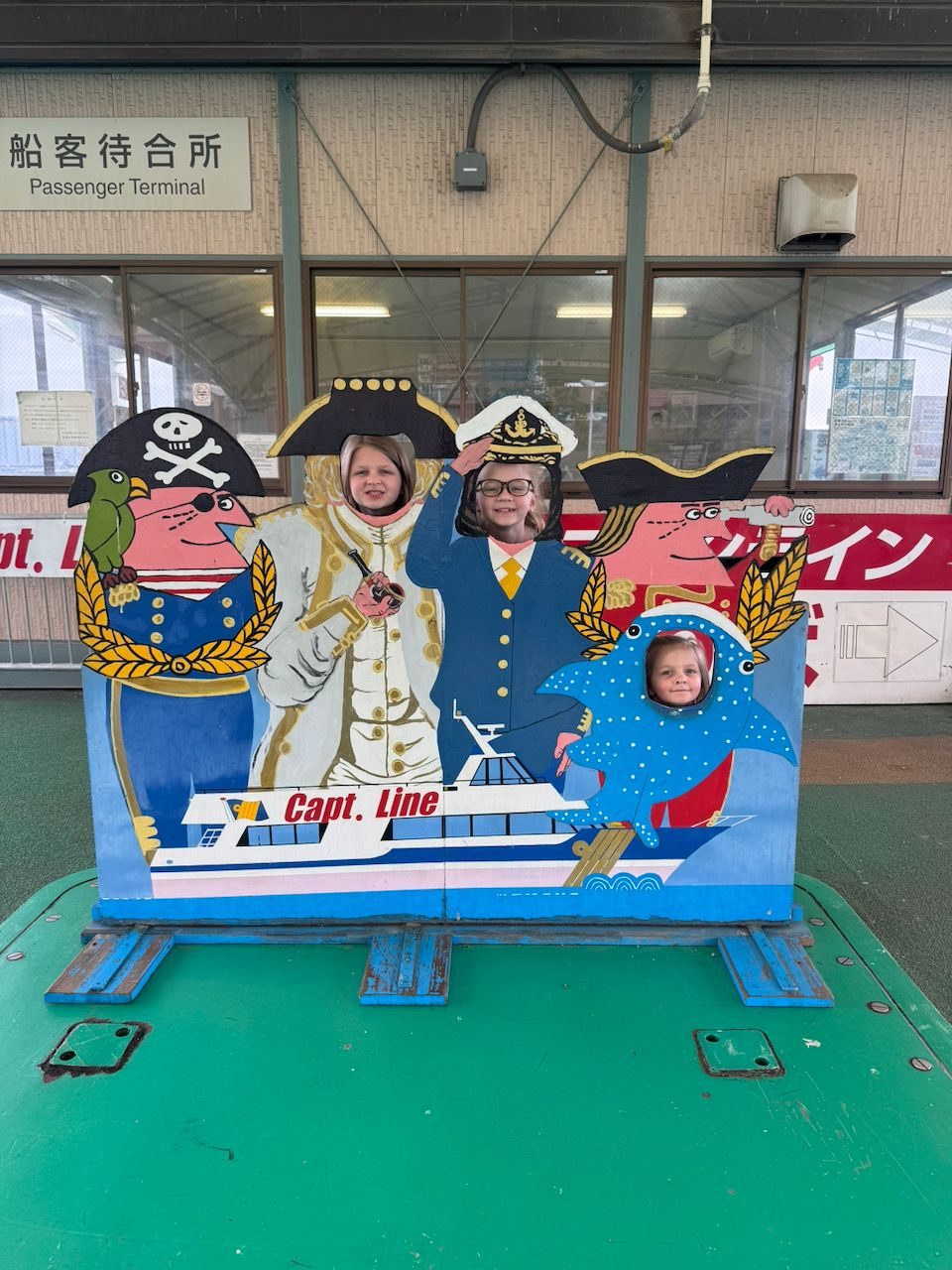 Three children pose with their faces through holes in a painted face-cutout board at a passenger ferry terminal, depicting nautical characters including a ship captain and a dolphin, with Japanese signage visible in the background.