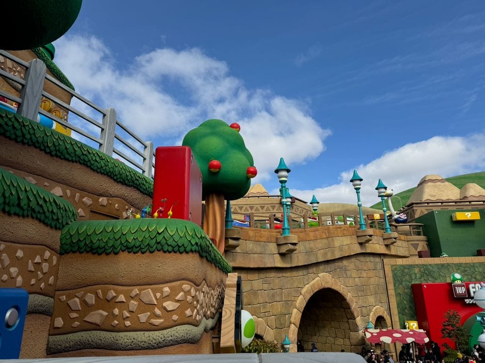 A low-angle view of the Super Nintendo World area at Universal Studios Japan, showing layered stone-and-grass terrain structures, a cartoonish green tree with red berries, teal lamp posts, a stone archway tunnel, and a 1-UP Factory shop sign under a blue sky with scattered clouds.