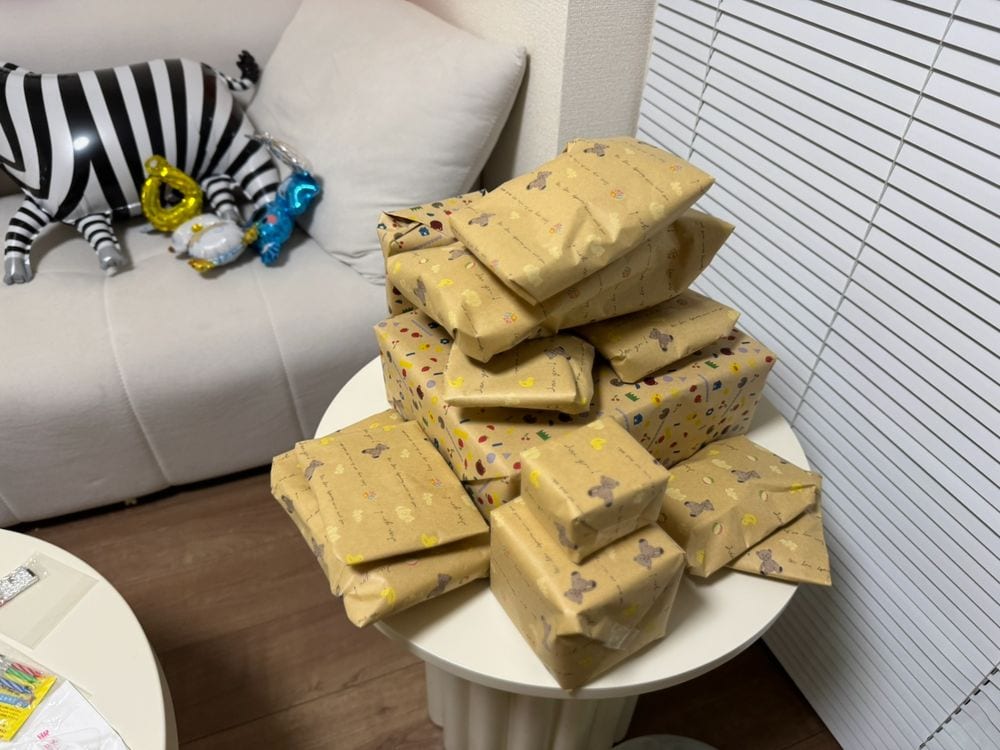 A small white side table stacked high with kraft paper-wrapped gifts in various sizes, with a black and white striped zebra balloon and colorful foil balloons visible on a white sofa in the background of a Japanese apartment.
