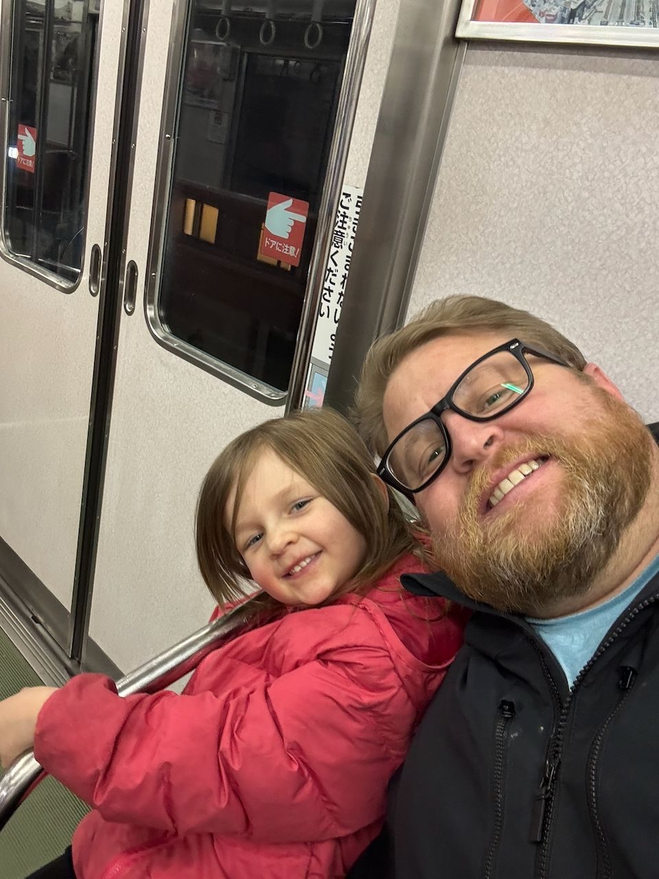 A man with glasses and a red beard takes a selfie with a young girl in a red puffer jacket on a Japanese subway car, both smiling at the camera.