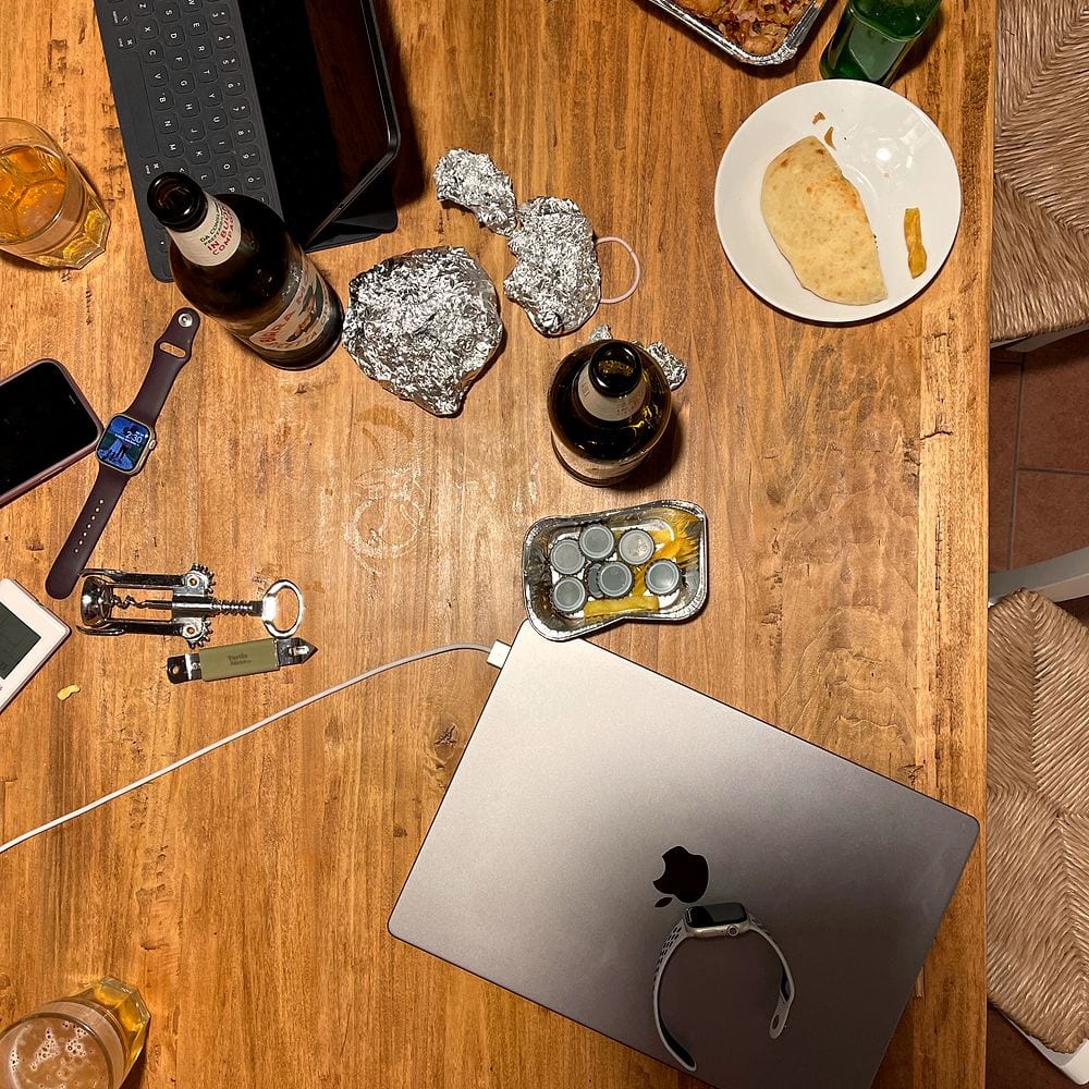 Overhead shot of a wooden farmhouse table with two beer bottles, crumpled foil, a plate with leftover flatbread and a candle, late at night in a Tuscan apartment