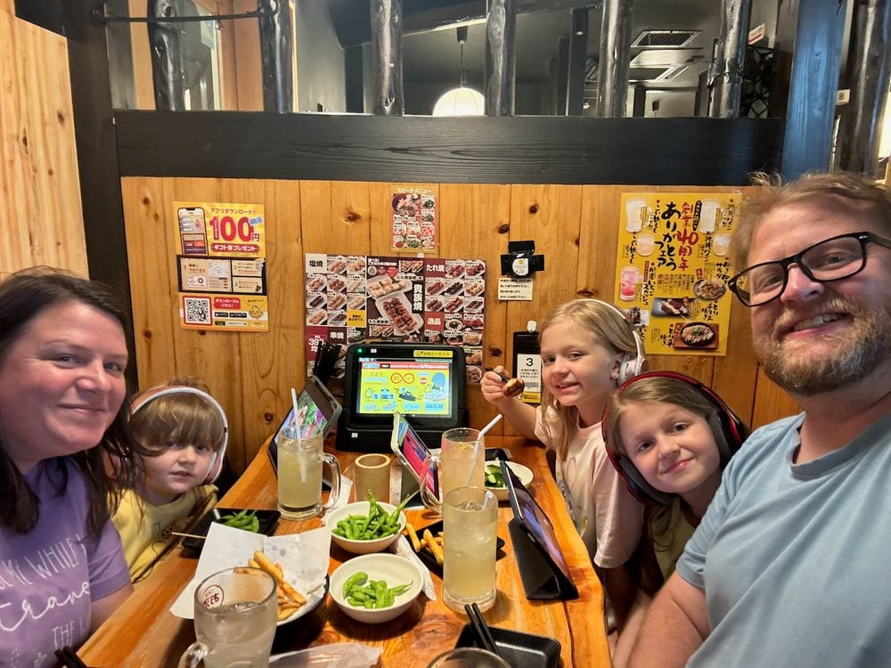 Family of five smiling at a yakitori restaurant booth in Asakusa with edamame and lemonade on the table