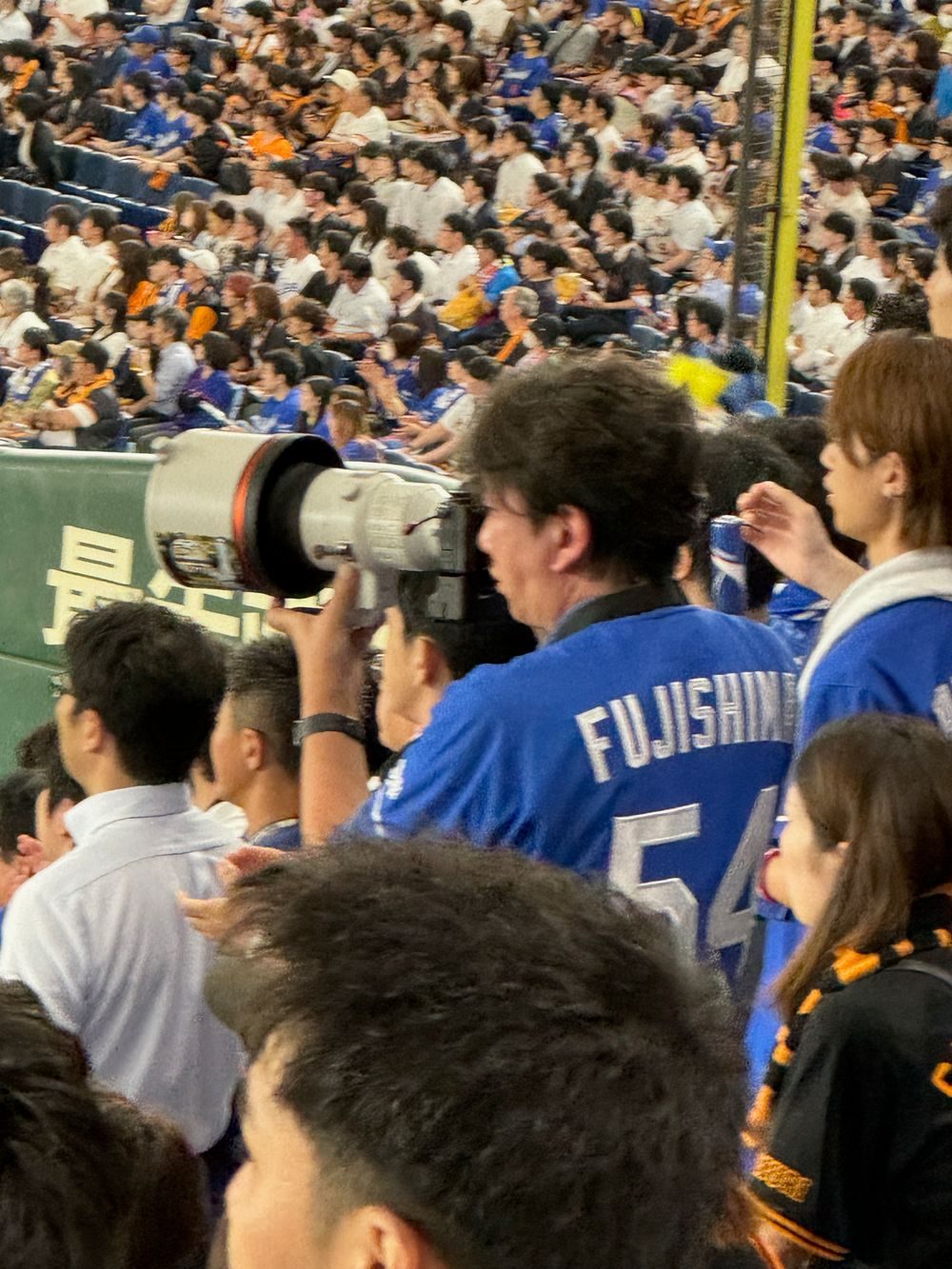 A fan in a blue Chunichi Dragons jersey with the name Fujishima and number 54 holds up a large camera with a telephoto lens at Tokyo Dome.