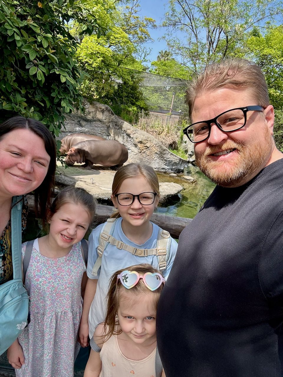 A family of five takes a selfie in front of a hippo enclosure at a zoo in Osaka, with two hippos resting near water in the background and the youngest child wearing pink heart-shaped sunglasses.