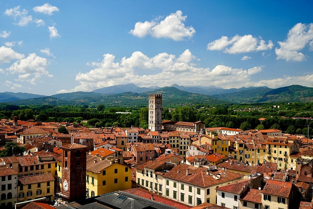 Wide aerial view over Lucca from the top of Guinigi Tower, showing terracotta rooftops and colourful buildings with a medieval tower rising in the centre