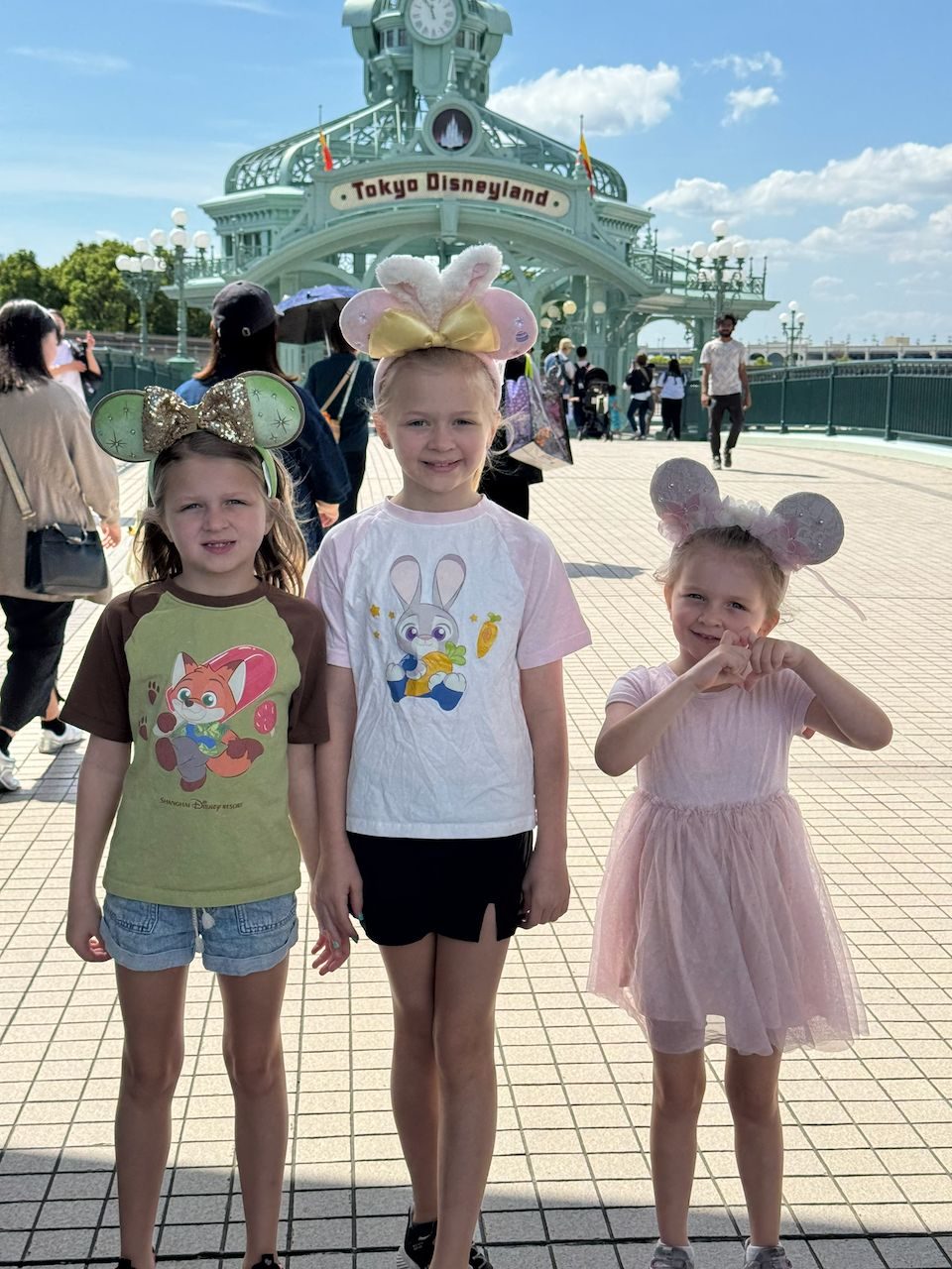 Three young girls wearing Minnie Mouse ears stand smiling in front of the Tokyo Disneyland entrance gate on a sunny day, each wearing Disney-themed shirts and casual outfits.