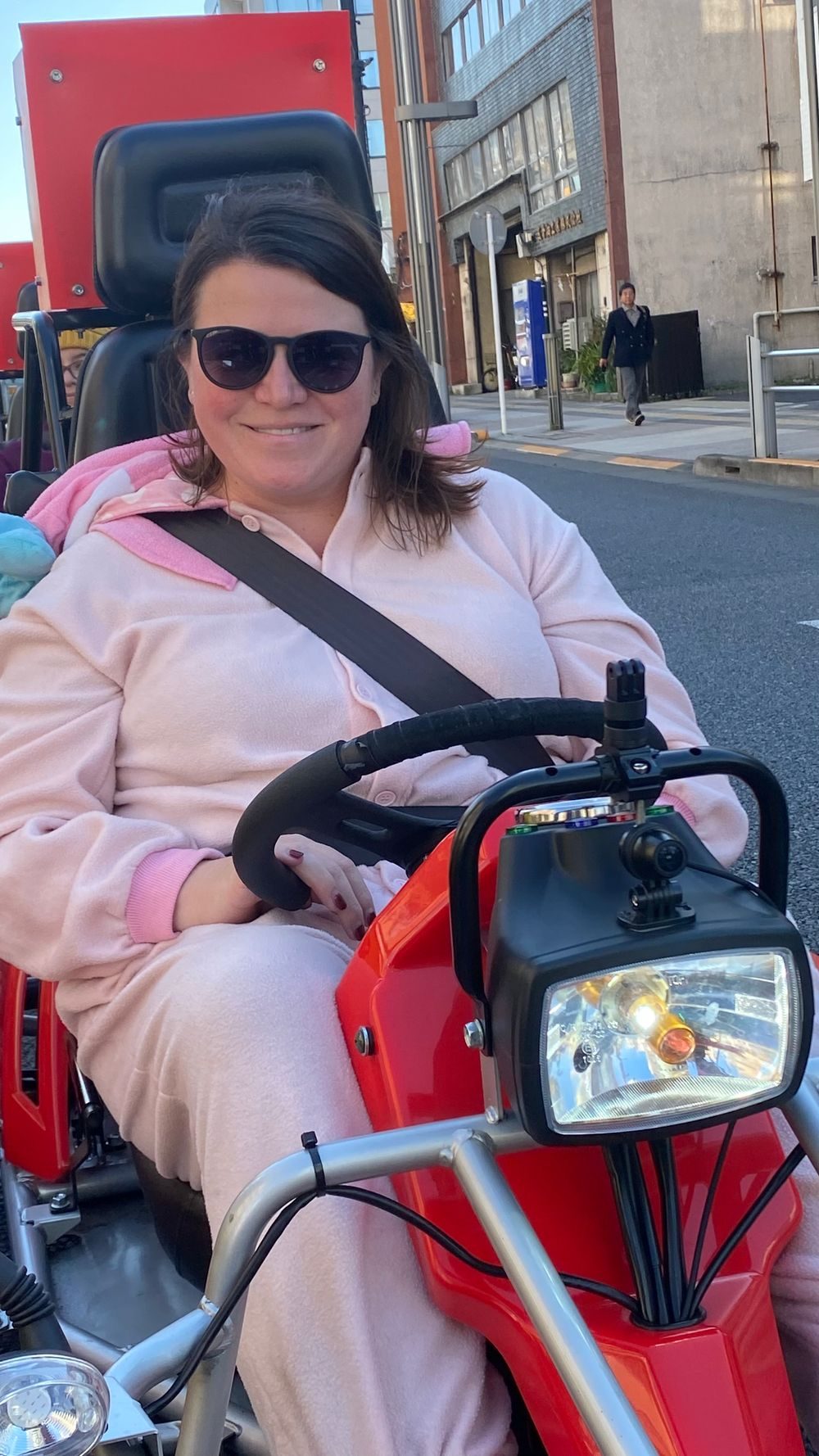 Lindsay, wearing a pink My Melody costume and sunglasses, sits behind the wheel of a red go-kart on a Tokyo street, smiling at the camera.