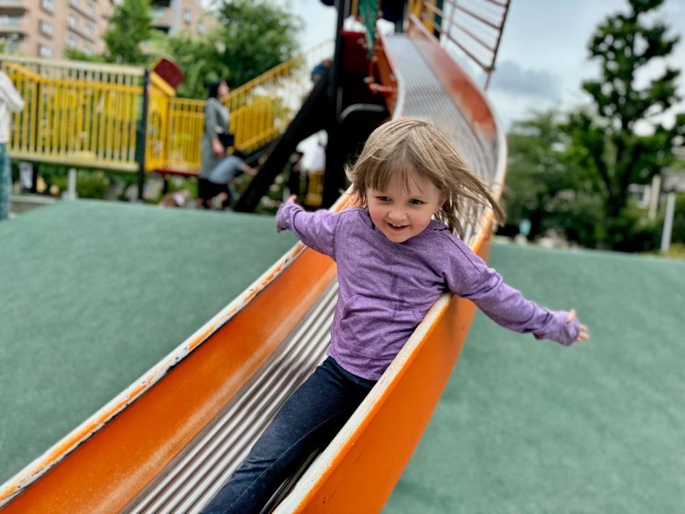 A young girl slides down a wide orange slide at Iriya Minami Park in Tokyo, arms outstretched and smiling, with playground structures visible in the background.
