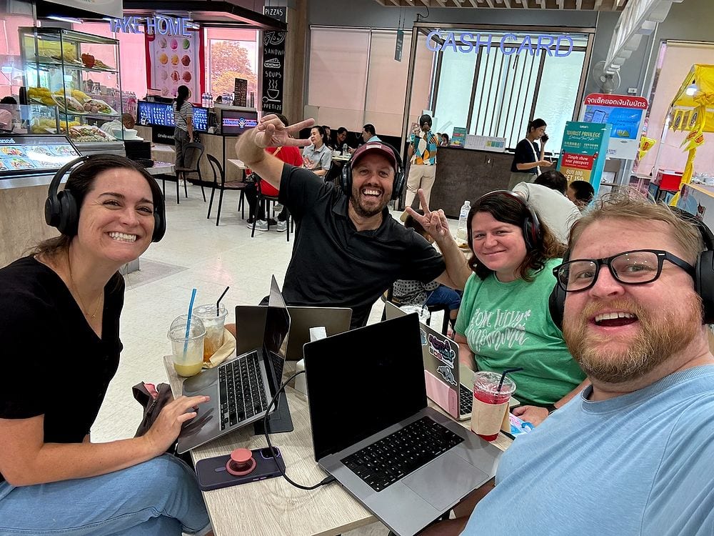 Four adults wearing headphones sit around a table covered in laptops and iced drinks in a busy Thai mall food court, grinning at the camera