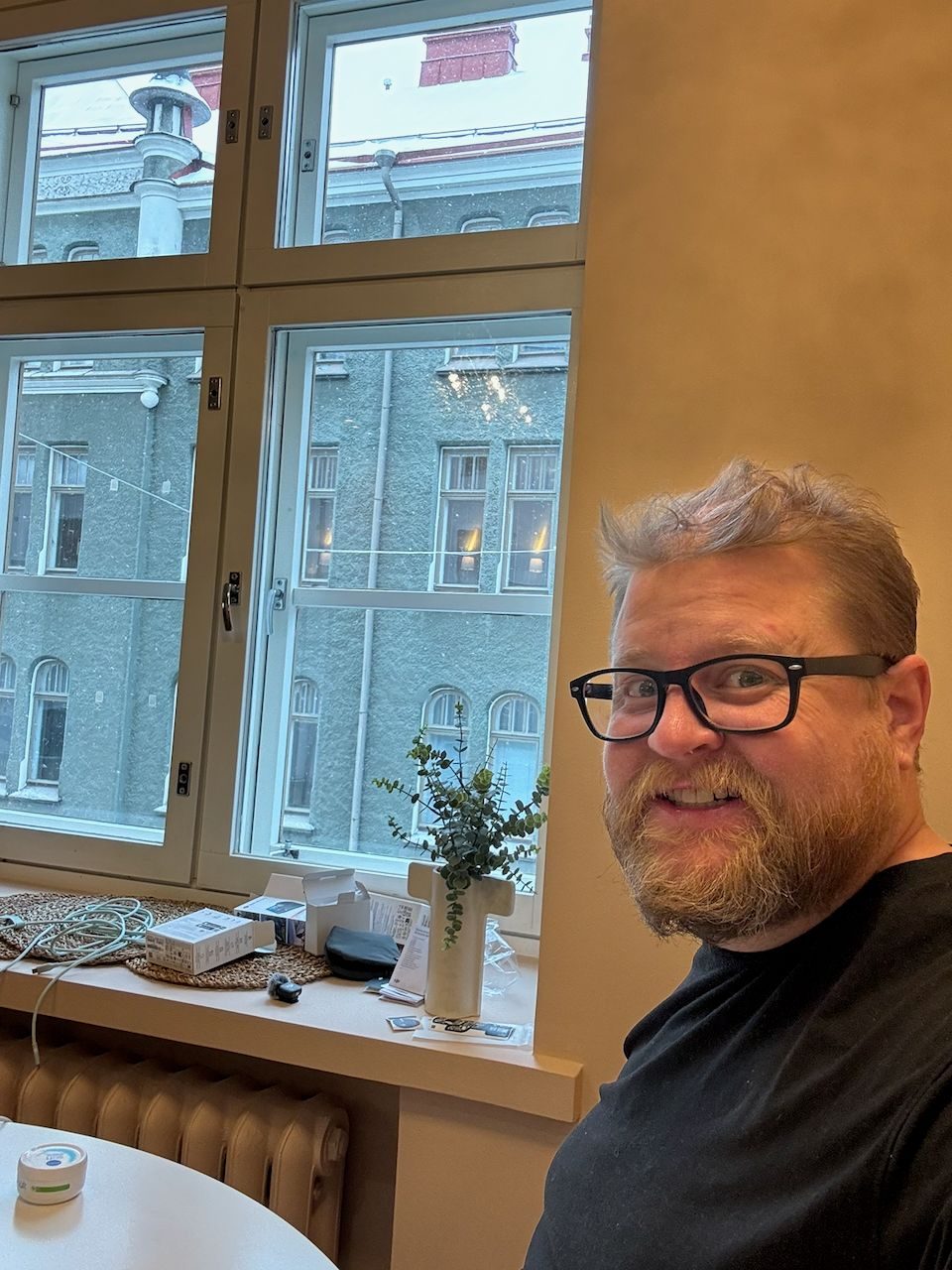 A smiling bearded man with glasses takes a selfie inside a cozy apartment, with a snow-dusted Helsinki street and a historic stone building visible through the window behind him