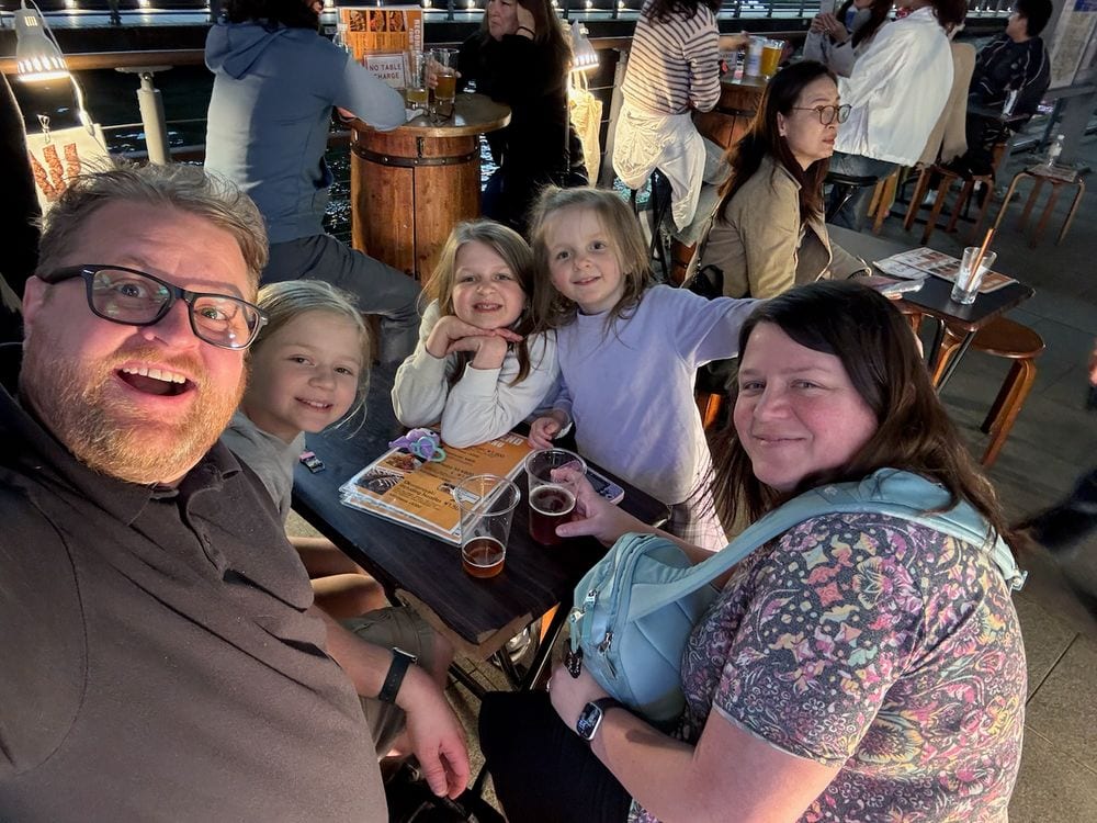 A family of five at an outdoor riverside bar table at night in Osaka, with two adults on either side, three young girls in the middle, and beers on the table.