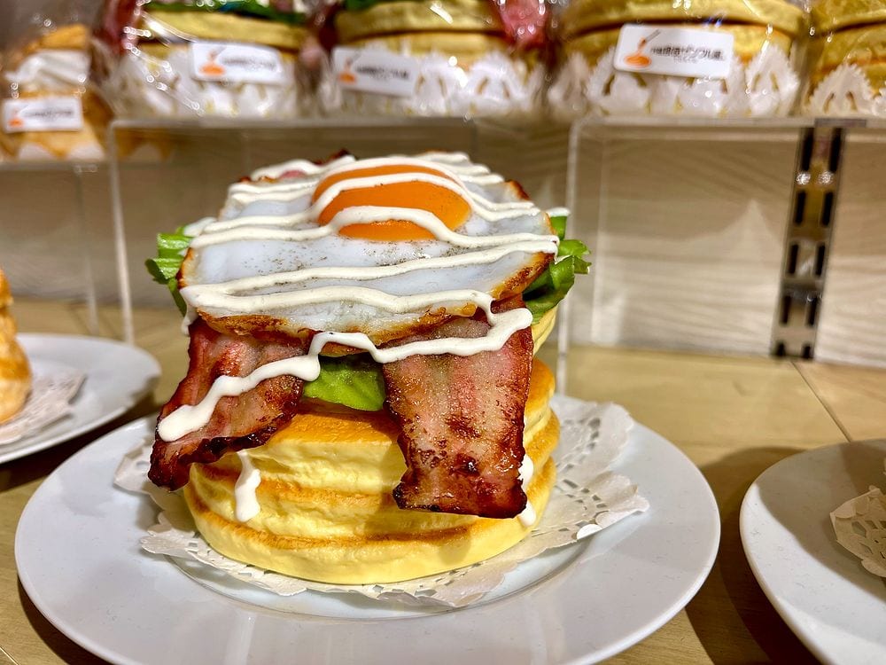 A hyper-realistic plastic food replica of a stacked pancake sandwich with bacon, lettuce, and a fried egg, displayed on a plate in a Kappabashi shop in Tokyo.