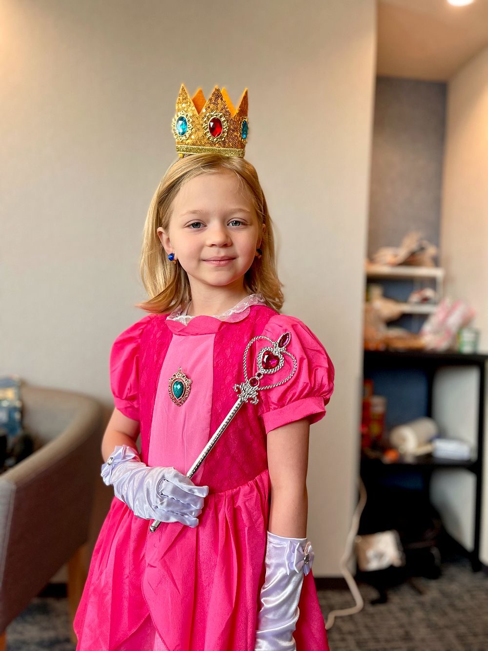 A young girl wears a pink Princess Peach costume with a jeweled crown, pink gloves, and a small scepter.