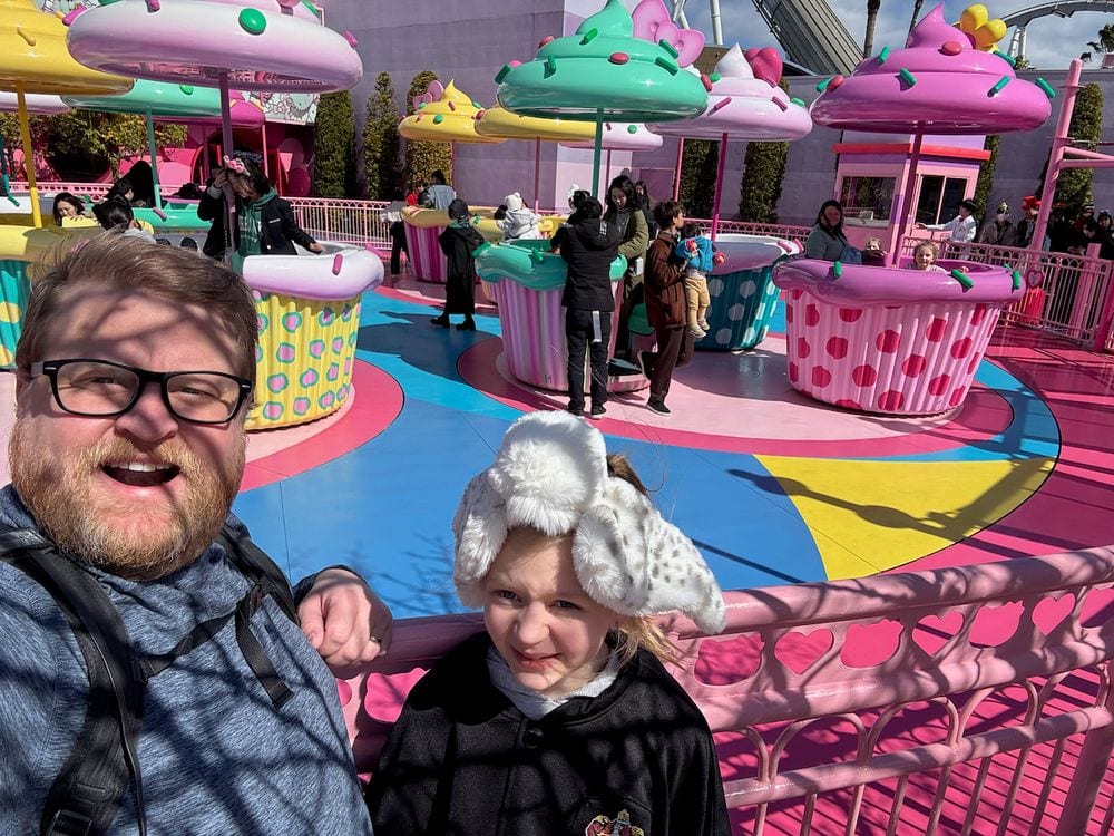 Adam and Lily take a selfie in front of the Hello Kitty cupcake spinning ride at Universal Studios Japan, surrounded by bright pink, yellow, and teal theming, with Lily wearing a white fluffy ear hat and making a serious face.