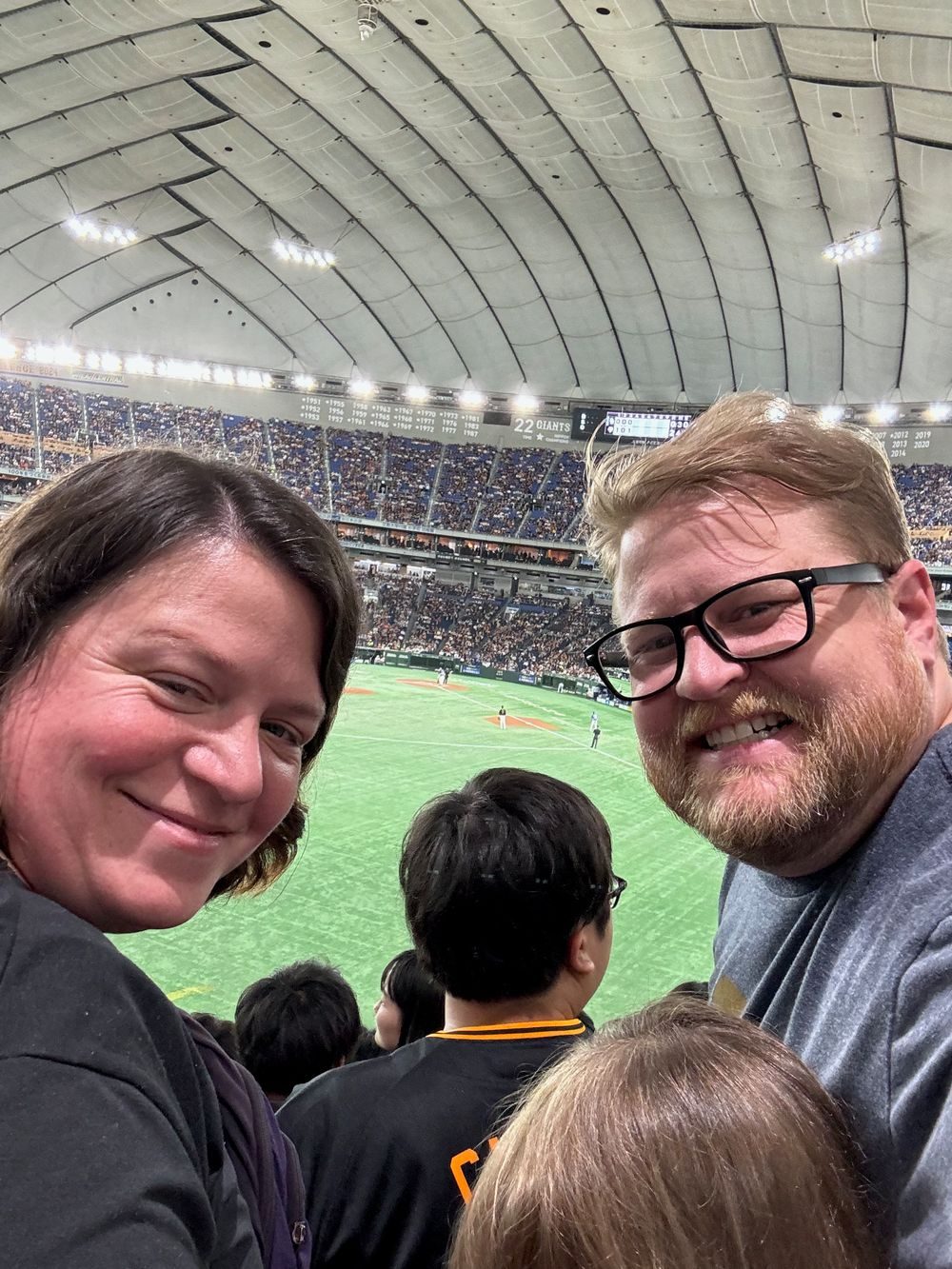 A man and woman smile together with the Tokyo Dome field and crowd behind them.