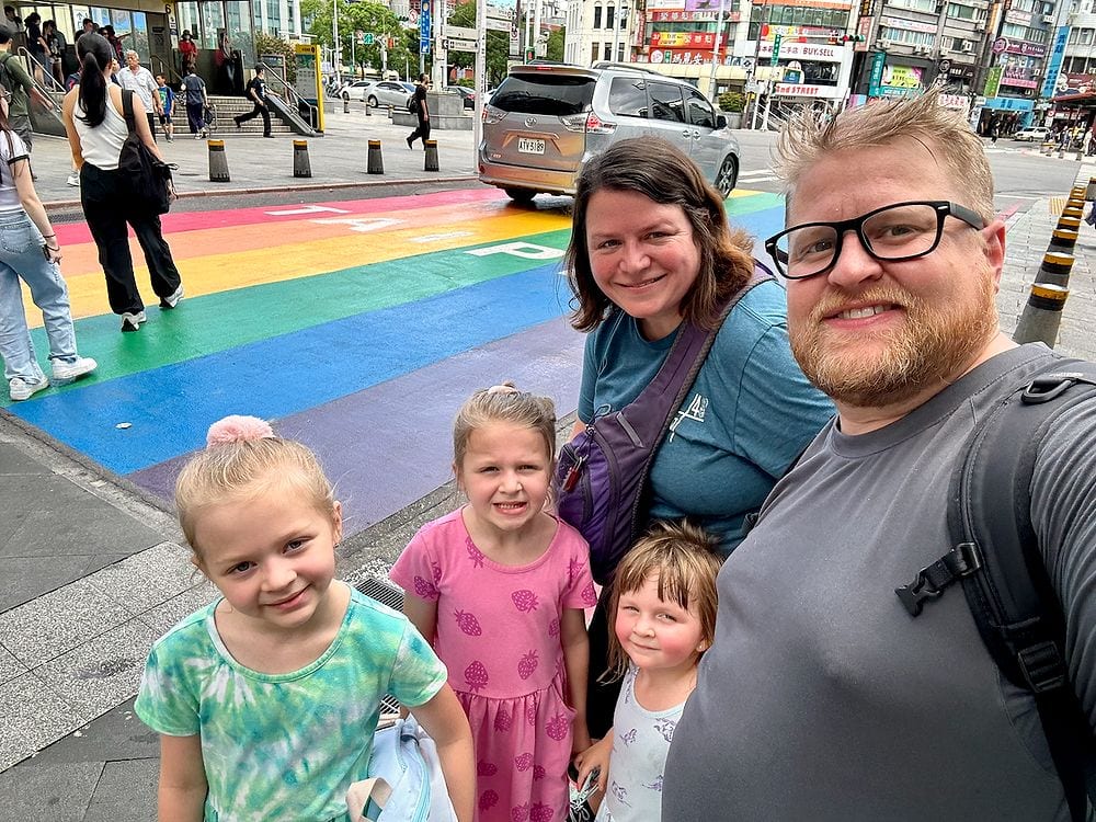 A family selfie taken on a busy Taipei sidewalk in front of a large rainbow-painted crosswalk, with two adults and three young girls smiling at the camera and city streets visible behind them.