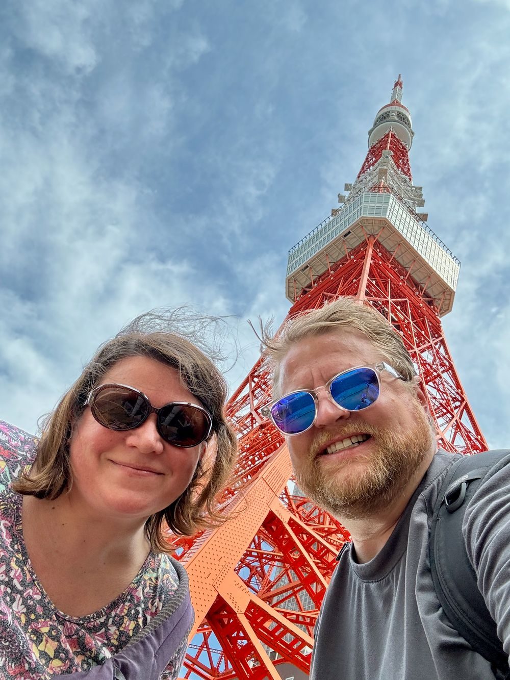 Adam and Lindsay take a selfie from directly below Tokyo Tower, the red lattice structure rising into a partly cloudy sky behind them, both wearing sunglasses.