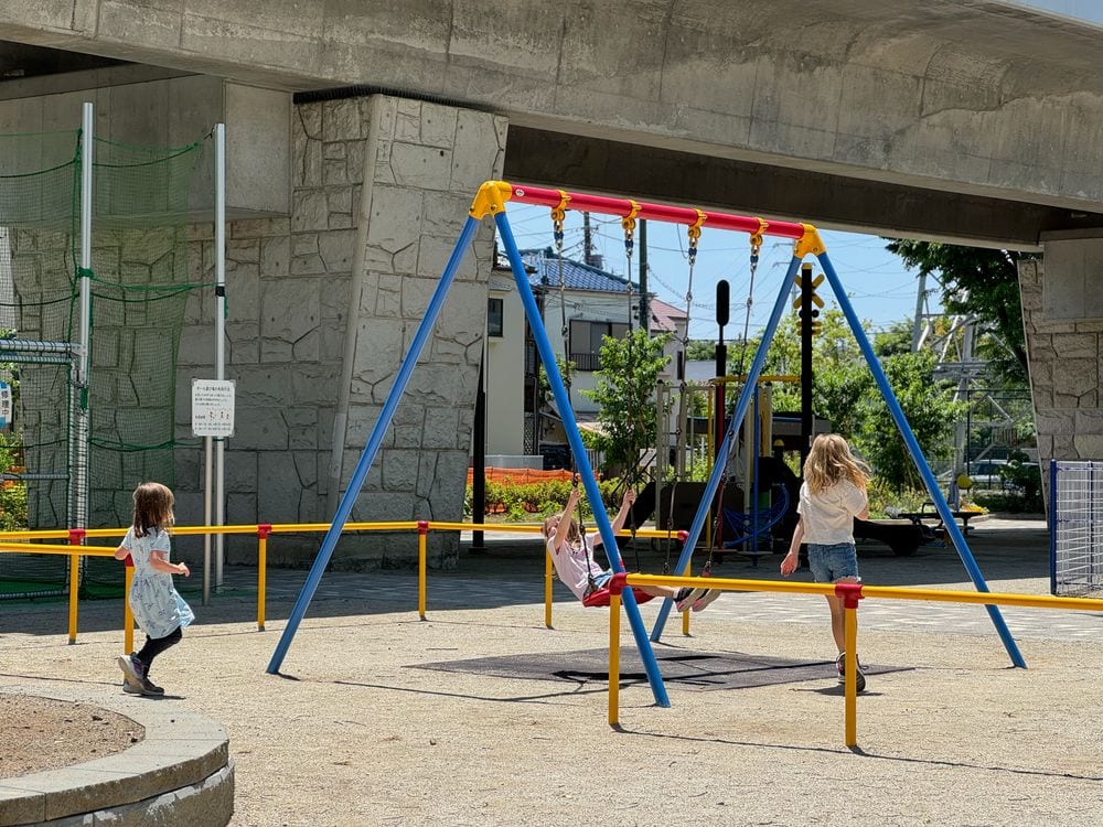 A wide shot of a swing set at a neighborhood playground in Tokyo, tucked under an elevated train line, with a girl walking toward it.