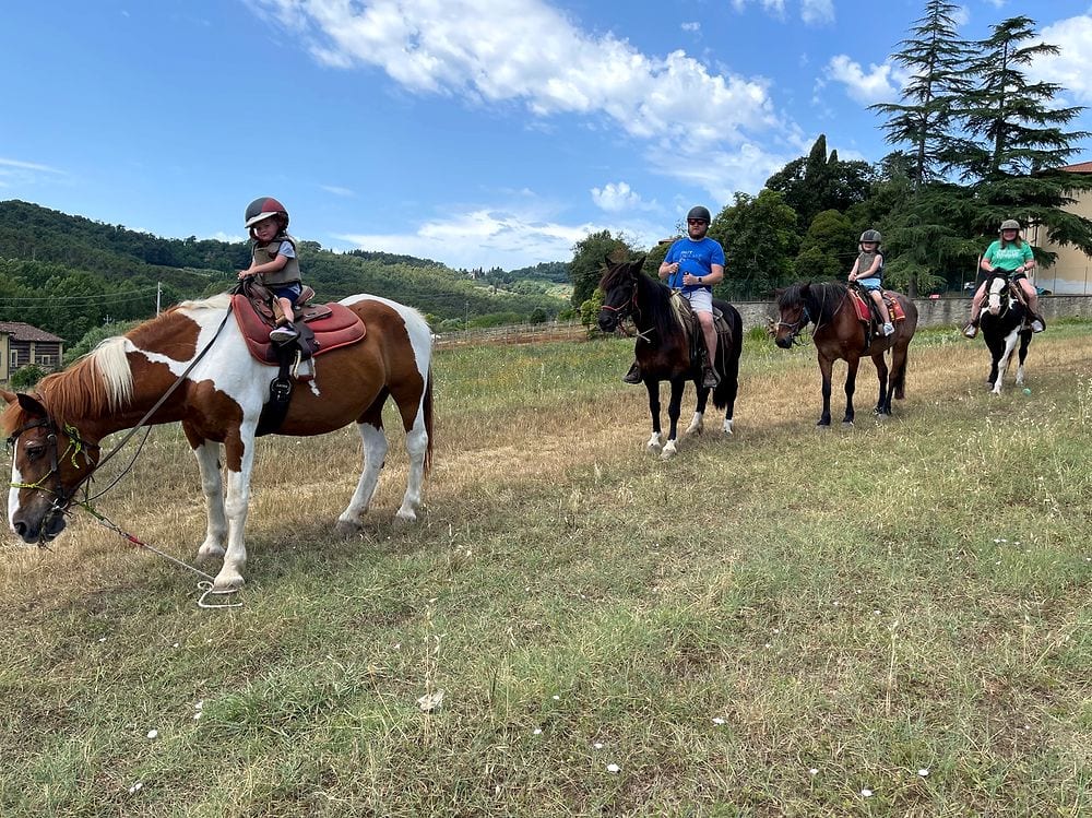 Four riders on horses and ponies lined up across a dry grassy field in the Tuscan hills near Lucca, all wearing helmets and protective vests