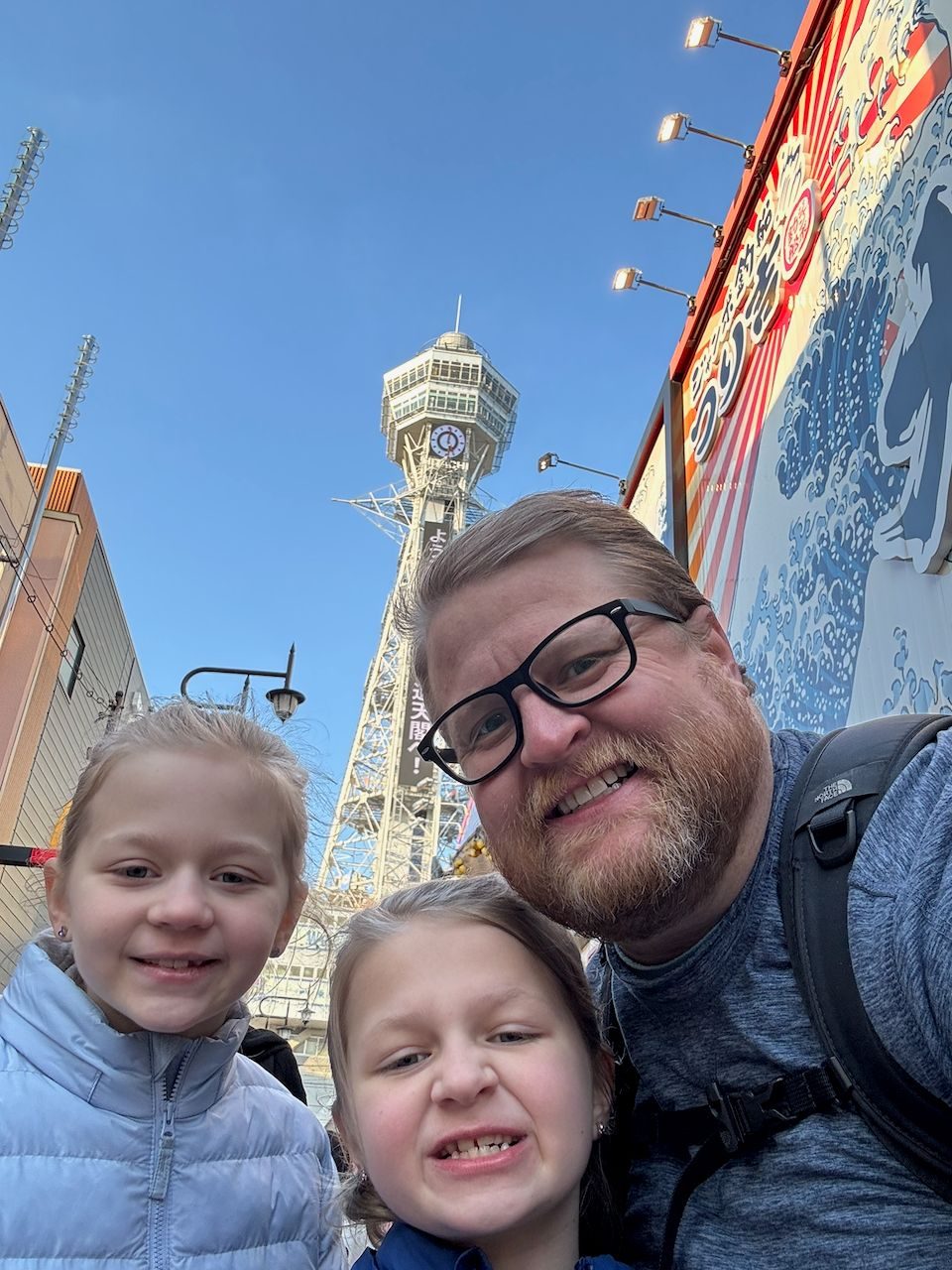 Adam takes a selfie with Lily and Cora in front of Tsutenkaku tower in Osaka's Shinsekai district on a clear blue-sky day, with a large Japanese advertisement sign visible to the right.