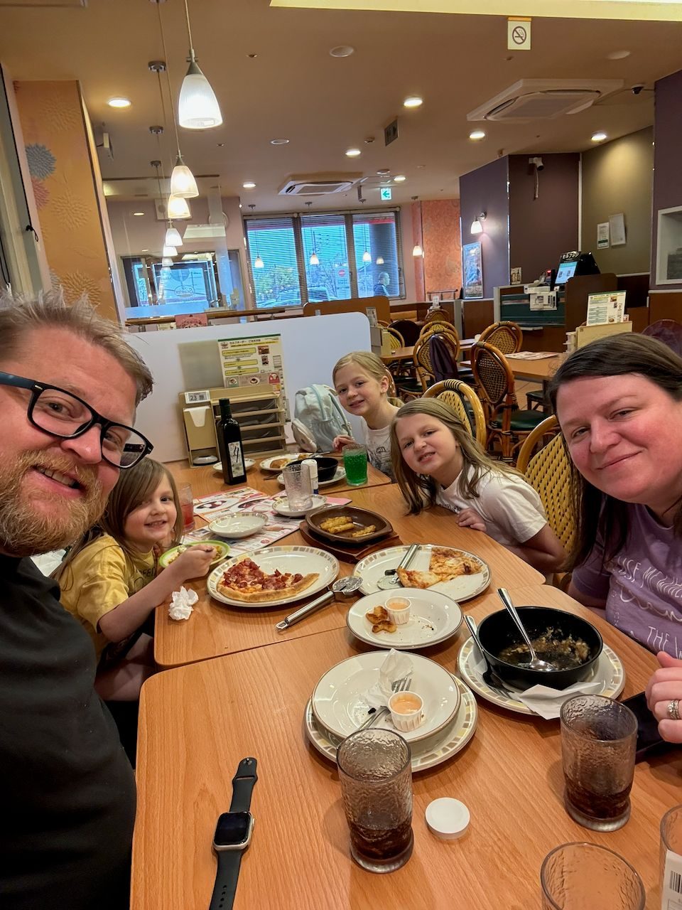 Adam takes a selfie at a restaurant table in Osaka with Lindsay, and their three daughters, plates of pizza and other dishes spread across the table in front of them.