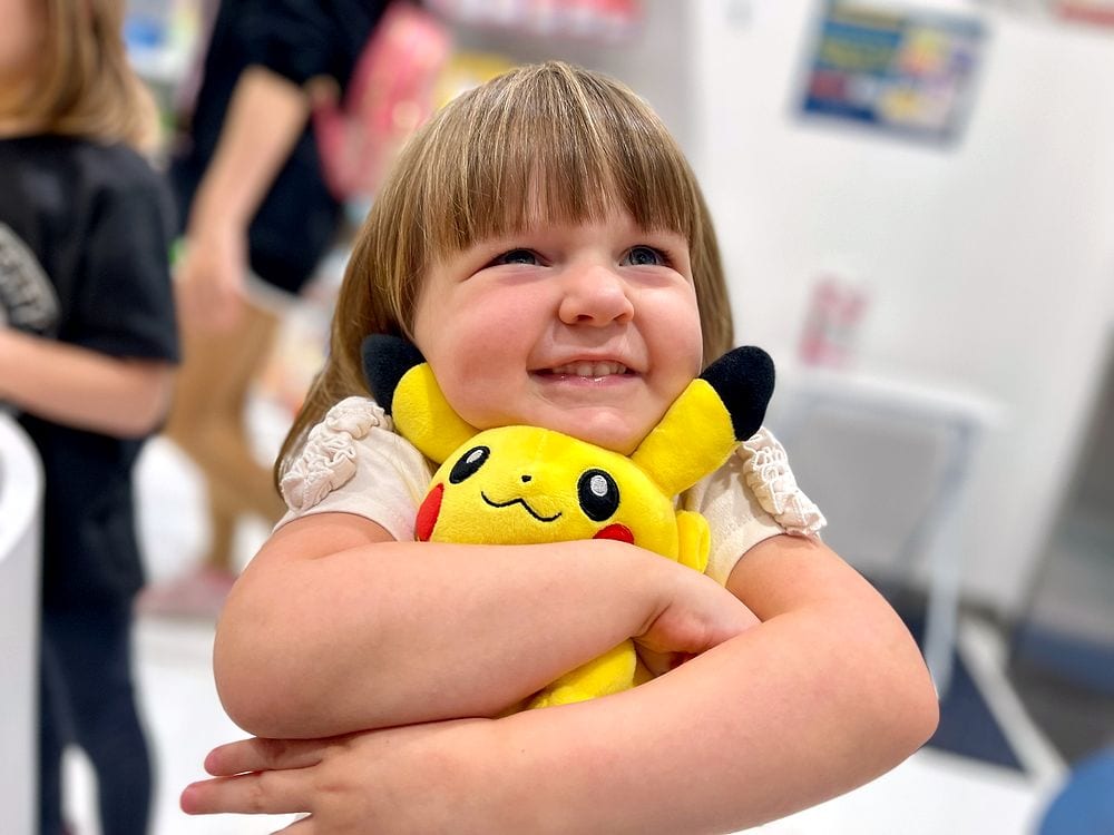 A young girl smiles while hugging a yellow Pikachu plush toy inside a brightly lit store.
