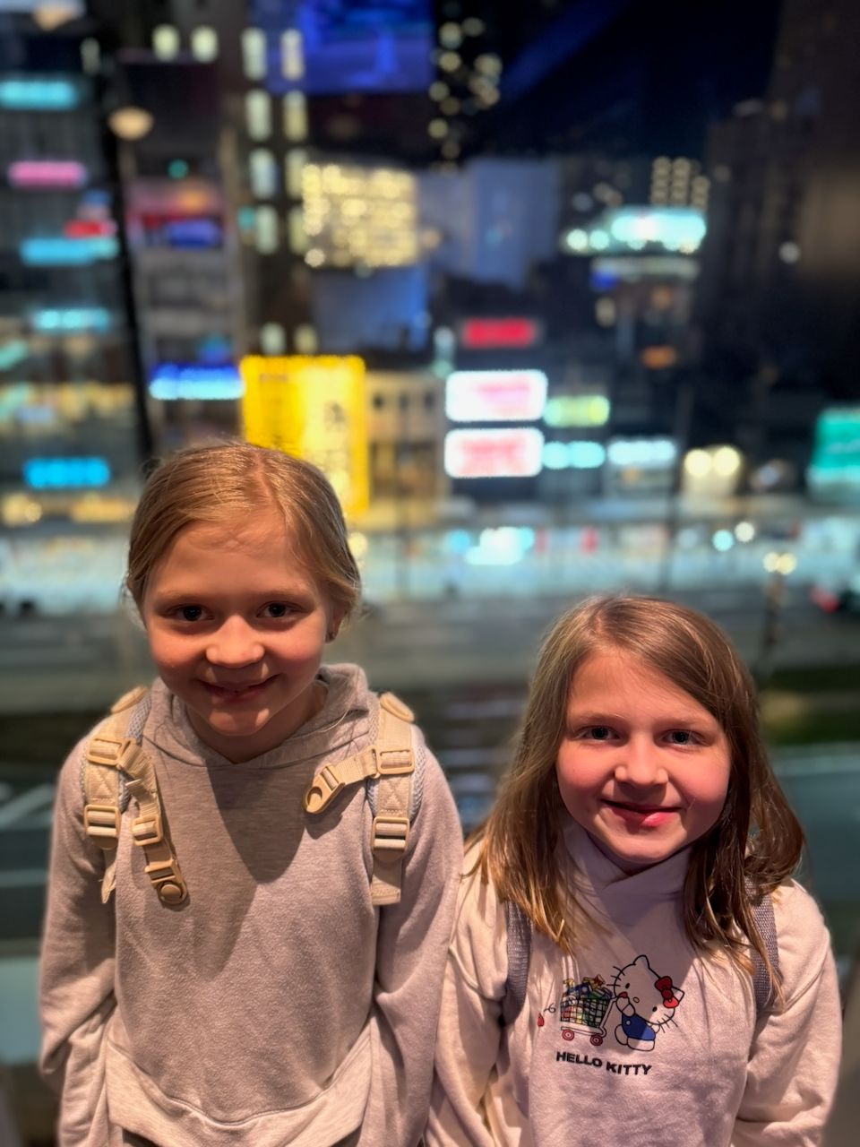 Two young girls wearing backpacks smile at the camera with the glowing neon signs of Osaka's Abenosuji district blurred in the background at night.