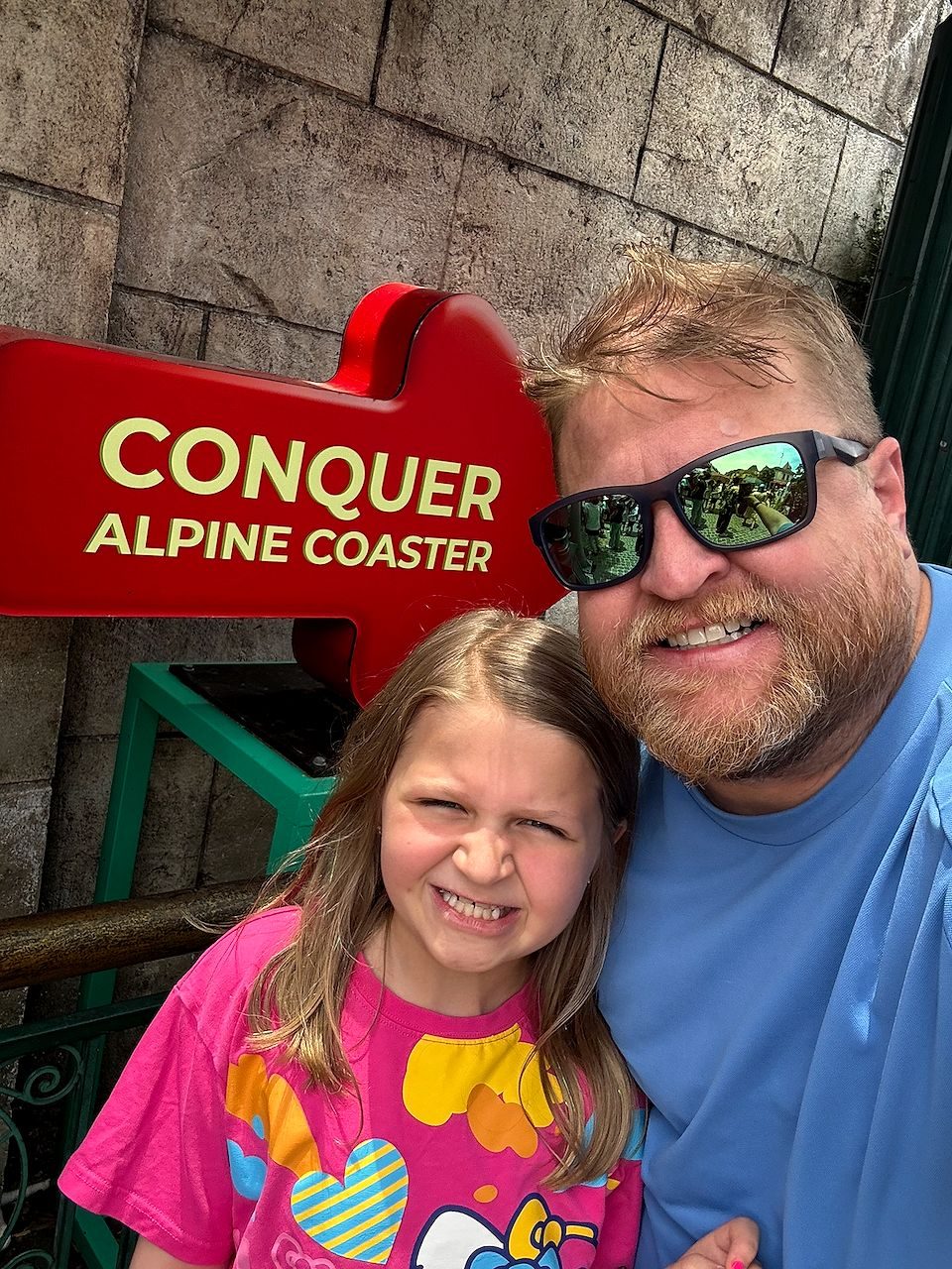 A man in sunglasses and a blue shirt takes a selfie with a young girl in a pink floral shirt in front of a red arrow sign reading Conquer Alpine Coaster, the girl making a fierce squinting expression.