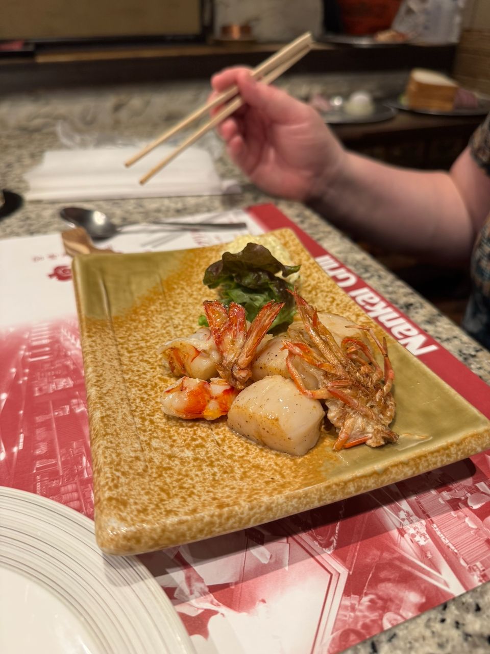 A ceramic plate of seared scallops and whole shrimp with greens on a restaurant table, with a hand holding chopsticks visible in the background.